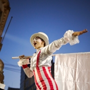 Fotografia realizada ao ar livre ao entardecer mostra uma pessoa caracterizada para uma apresenta&ccedil;&atilde;o art&iacute;stica em espa&ccedil;o urbano. A pessoa veste roupa branca com detalhes vermelhos, incluindo cal&ccedil;as listradas, suspens&oacute;rios e chap&eacute;u claro. Com um dos bra&ccedil;os estendido para a frente, segura sobre os ombros uma haste da qual pende um grande tecido branco. A cena &eacute; registrada em &acirc;ngulo baixo, destacando o c&eacute;u azul ao fundo e parte da fachada de um pr&eacute;dio hist&oacute;rico com sacada e elementos ornamentais. A ilumina&ccedil;&atilde;o valoriza o contraste entre o figurino claro e o ambiente urbano ao redor.