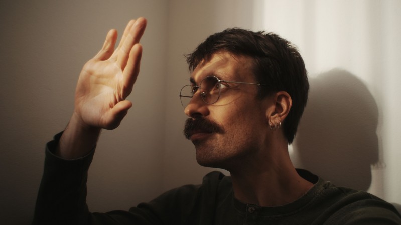 A imagem mostra o cantor Pedro Longes de perfil, posicionado do lado direito da composi&ccedil;&atilde;o, voltada para a esquerda. Ela tem cabelos curtos e escuros, usa &oacute;culos de arma&ccedil;&atilde;o fina e arredondada, um brinco de argola pequeno na orelha esquerda e um bigode espesso. Veste uma blusa de manga comprida em tom escuro. O bra&ccedil;o esquerdo est&aacute; levantado, com a m&atilde;o aberta e os dedos estendidos, como se tentasse tocar ou bloquear a luz que incide sobre ela. Um feixe de luz quente entra pela direita da cena, iluminando parte do rosto, a m&atilde;o e o ombro, enquanto o restante permanece em sombras suaves. A ilumina&ccedil;&atilde;o tamb&eacute;m projeta a sombra n&iacute;tida da cabe&ccedil;a e da m&atilde;o sobre a parede clara ao fundo.