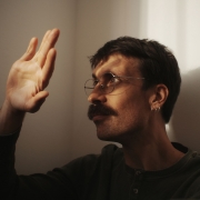 A imagem mostra o cantor Pedro Longes de perfil, posicionado do lado direito da composi&ccedil;&atilde;o, voltada para a esquerda. Ela tem cabelos curtos e escuros, usa &oacute;culos de arma&ccedil;&atilde;o fina e arredondada, um brinco de argola pequeno na orelha esquerda e um bigode espesso. Veste uma blusa de manga comprida em tom escuro. O bra&ccedil;o esquerdo est&aacute; levantado, com a m&atilde;o aberta e os dedos estendidos, como se tentasse tocar ou bloquear a luz que incide sobre ela. Um feixe de luz quente entra pela direita da cena, iluminando parte do rosto, a m&atilde;o e o ombro, enquanto o restante permanece em sombras suaves. A ilumina&ccedil;&atilde;o tamb&eacute;m projeta a sombra n&iacute;tida da cabe&ccedil;a e da m&atilde;o sobre a parede clara ao fundo.