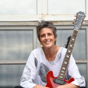 Fotografia mostra uma mulher sentada diante de uma estrutura de janelas com caixilhos metálicos claros. A pessoa veste roupa branca com estampas abstratas em preto e está apoiada sobre uma guitarra vermelha apoiada verticalmente ao lado do corpo. A composição é iluminada por luz natural suave. 