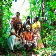 Cinco integrantes do Grupo Desagravo est&atilde;o reunidos em uma &aacute;rea de vegeta&ccedil;&atilde;o densa, cercados por &aacute;rvores, folhagens e plantas de diferentes alturas. Dois membros ocupam o primeiro plano em posi&ccedil;&atilde;o mais baixa, enquanto os demais aparecem atr&aacute;s, formando uma composi&ccedil;&atilde;o em diferentes n&iacute;veis. Os figurinos combinam tons terrosos, branco, amarelo e estampas variadas. A luz natural atravessa a copa das &aacute;rvores e cria contrastes de sombras e luminosidade sobre o grupo e a paisagem ao redor.