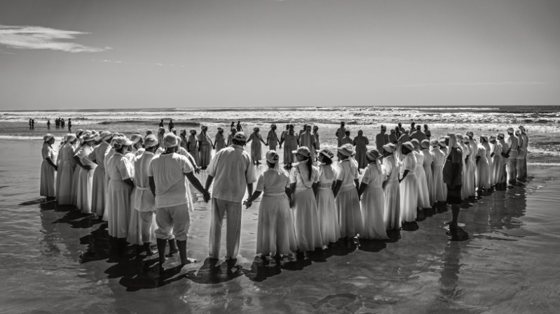 Fotografia em preto e branco mostra dezenas de pessoas reunidas em uma praia, formando um grande c&iacute;rculo de m&atilde;os dadas sobre a areia molhada. A maioria veste roupas claras e longas, que se destacam pelo contraste com o mar e o c&eacute;u ao fundo. O reflexo das figuras aparece na superf&iacute;cie &uacute;mida da praia, enquanto ondas se aproximam da faixa de areia. Outros grupos de pessoas podem ser vistos mais distantes, pr&oacute;ximos &agrave; &aacute;gua. A composi&ccedil;&atilde;o ampla enfatiza a forma&ccedil;&atilde;o circular e a intera&ccedil;&atilde;o coletiva diante da paisagem litor&acirc;nea.