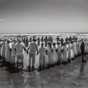 Fotografia em preto e branco mostra dezenas de pessoas reunidas em uma praia, formando um grande c&iacute;rculo de m&atilde;os dadas sobre a areia molhada. A maioria veste roupas claras e longas, que se destacam pelo contraste com o mar e o c&eacute;u ao fundo. O reflexo das figuras aparece na superf&iacute;cie &uacute;mida da praia, enquanto ondas se aproximam da faixa de areia. Outros grupos de pessoas podem ser vistos mais distantes, pr&oacute;ximos &agrave; &aacute;gua. A composi&ccedil;&atilde;o ampla enfatiza a forma&ccedil;&atilde;o circular e a intera&ccedil;&atilde;o coletiva diante da paisagem litor&acirc;nea.