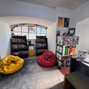 Fotografia mostra um espaço de leitura voltado para histórias em quadrinhos e literatura infantojuvenil. O ambiente tem paredes brancas, uma grande janela arqueada ao fundo e duas estantes baixas repletas de livros organizados lado a lado. No centro da sala há dois pufes grandes, um amarelo e um vermelho, posicionados sobre um tapete cinza. À direita, um expositor identificado com o texto “Gibiteca BPE” reúne capas de quadrinhos e ilustrações coloridas. Sobre o móvel e nas paredes próximas aparecem livros expostos, cartazes e pequenos objetos decorativos.