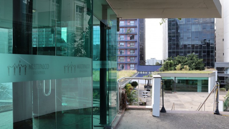 Fotografia mostra uma &aacute;rea externa do Multipalco Eva Sopher cercada por edif&iacute;cios urbanos. &Agrave; esquerda, uma fachada envidra&ccedil;ada exibe faixas com o logotipo e o nome &ldquo;Multipalco Eva Sopher&rdquo; aplicados sobre os pain&eacute;is de vidro. No centro da imagem, uma rampa e uma escadaria conduzem a um p&aacute;tio aberto com &aacute;reas ajardinadas e mobili&aacute;rio externo. Ao fundo, edif&iacute;cios residenciais e comerciais formam a paisagem urbana. A combina&ccedil;&atilde;o entre estruturas de vidro, concreto e &aacute;reas verdes destaca a arquitetura contempor&acirc;nea do complexo cultural.