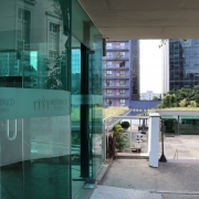 Fotografia mostra uma &aacute;rea externa do Multipalco Eva Sopher cercada por edif&iacute;cios urbanos. &Agrave; esquerda, uma fachada envidra&ccedil;ada exibe faixas com o logotipo e o nome &ldquo;Multipalco Eva Sopher&rdquo; aplicados sobre os pain&eacute;is de vidro. No centro da imagem, uma rampa e uma escadaria conduzem a um p&aacute;tio aberto com &aacute;reas ajardinadas e mobili&aacute;rio externo. Ao fundo, edif&iacute;cios residenciais e comerciais formam a paisagem urbana. A combina&ccedil;&atilde;o entre estruturas de vidro, concreto e &aacute;reas verdes destaca a arquitetura contempor&acirc;nea do complexo cultural.