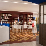 Fotografia mostra a entrada de uma biblioteca ou sala de leitura vista através de amplas portas de madeira abertas. No interior, estantes altas em tom vinho ocupam o centro do ambiente e estão repletas de livros. Mesas brancas com cadeiras do mesmo tom formam uma área de leitura iluminada por luminárias de mesa. O piso apresenta um padrão geométrico em tons de amarelo e vermelho, enquanto detalhes arquitetônicos azuis contornam parte do teto e das paredes. À direita, uma pessoa em movimento atravessa o espaço, levemente desfocada, reforçando a atmosfera de uso e circulação do local.