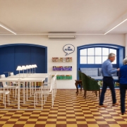 Fotografia de uma biblioteca ou espaço de leitura amplo e iluminado. À esquerda, grandes estantes repletas de livros ocupam a parede e contêm uma sinalização com a palavra “Manga”. No centro, mesas brancas organizadas em conjunto são iluminadas por luminárias alinhadas. Ao fundo, nichos azuis embutidos nas paredes servem como áreas de permanência e leitura. Duas pessoas caminham pelo ambiente e aparecem levemente desfocadas pelo movimento. O piso é quadriculado em tons de amarelo e vermelho.