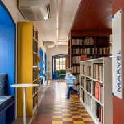 Fotografia de um corredor de biblioteca cercado por estantes cheias de livros dos dois lados. À esquerda, há um nicho de leitura azul com almofada e uma pequena mesa circular branca. O piso em padrão geométrico amarelo e vermelho conduz o olhar até uma janela ao fundo, emoldurada por detalhes azuis. No centro da imagem, uma criança em movimento cruza o corredor, aparecendo desfocada devido ao deslocamento. Em uma das estantes, destaca-se uma sinalização vertical com a palavra “Marvel”.
