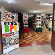 Fotografia registra a entrada de uma gibiteca vista a partir de uma porta aberta. Em primeiro plano, um grande painel exibe o nome “Gibiteca BPE” sobre uma composição formada por capas de histórias em quadrinhos. O interior do espaço revela longas estantes repletas de livros e gibis distribuídas ao longo de um corredor amplo. O piso apresenta um padrão quadriculado em tons de amarelo e vermelho, enquanto bancos estofados verdes e vermelhos estão espalhados pelo ambiente para leitura e permanência. A perspectiva da imagem conduz o olhar até o fundo da biblioteca, evidenciando a extensão do acervo.