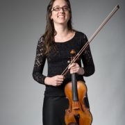 A imagem mostra uma mulher adulta, a violinista Brigitta Calloni. Ela tem cabelos longos e castanhos, usa óculos e sorri para a câmera. A instrumentista veste uma blusa preta de mangas longas com detalhes rendados e uma saia preta. A mulher segura um violino de madeira alaranjada à frente do corpo e, com a outra mão, segura o arco do instrumento, que está inclinado para o alto à direita. O fundo é liso, em tom cinza, e a iluminação destaca a mulher e o instrumento.