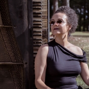 A imagem mostra a pianista Catarina Domenici. Ela tem cabelos curtos e crespos, usa óculos escuros e brincos. A mulher veste uma blusa preta de um ombro só e está sentada ao lado de um grande piano antigo, cuja estrutura de madeira escura e parte das teclas aparecem à esquerda da imagem. Ela olha para o lado, com uma expressão tranquila. Ao fundo, há árvores e vegetação, indicando um ambiente ao ar livre, com luz natural incidindo sobre a cena.