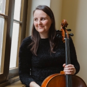 A imagem mostra Martina Stroher, uma mulher adulta que está posicionada ao lado de uma janela alta com estrutura de madeira e vidros divididos em painéis. Ela segura um instrumento de cordas de grande porte, aparentando ser um violoncelo, apoiado verticalmente junto ao corpo. Ela tem cabelos longos, escuros e estão soltos. A pessoa veste uma blusa de manga comprida de cor preta e textura felpuda. Uma das mãos segura a região superior do violoncelo, próxima às cravelhas, enquanto a outra está apoiada sobre a parte frontal do instrumento.