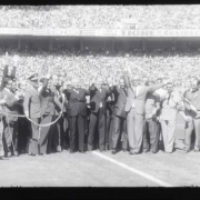 Fotograma em preto e branco registra uma cerimônia realizada dentro de um estádio lotado. No centro da imagem, diversas pessoas estão reunidas sobre o gramado, algumas com os braços erguidos, enquanto um grupo segura uma longa fita, sugerindo um ato de inauguração ou celebração oficial. Ao fundo, as arquibancadas aparecem completamente ocupadas pelo público. A imagem apresenta granulação, bordas escuras e características típicas de registros cinematográficos históricos.