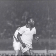 Fotografia em preto e branco presente no documentário “Cinejornal Gigante da Beira-Rio – Inauguração do Estádio José Pinheiro Borda”. Mostra o jogador Pelé em primeiro plano, ele é um homem negro e utiliza uma camisa clara da seleção brasileira. Ao fundo, outro atleta aparece desfocado, enquanto a arquibancada e o restante do estádio ficam fora de foco. Marcas e riscos visíveis na imagem revelam o aspecto de filme antigo.