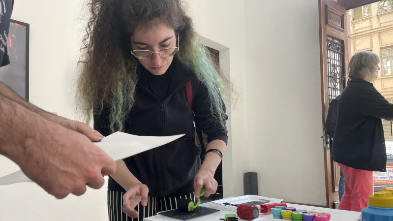 Fotografia mostra uma atividade pr&aacute;tica de impress&atilde;o artesanal realizada em um espa&ccedil;o expositivo ou educativo. Em primeiro plano, uma pessoa utiliza um pequeno rolo para aplicar tinta sobre uma matriz apoiada em uma mesa. Ao lado est&atilde;o organizados recipientes com tintas coloridas, bandejas, carimbos e outros materiais de impress&atilde;o. Outra pessoa segura uma folha de papel pr&oacute;xima &agrave; &aacute;rea de trabalho. Ao fundo, uma porta aberta permite a entrada de luz natural e revela parte da arquitetura do pr&eacute;dio.