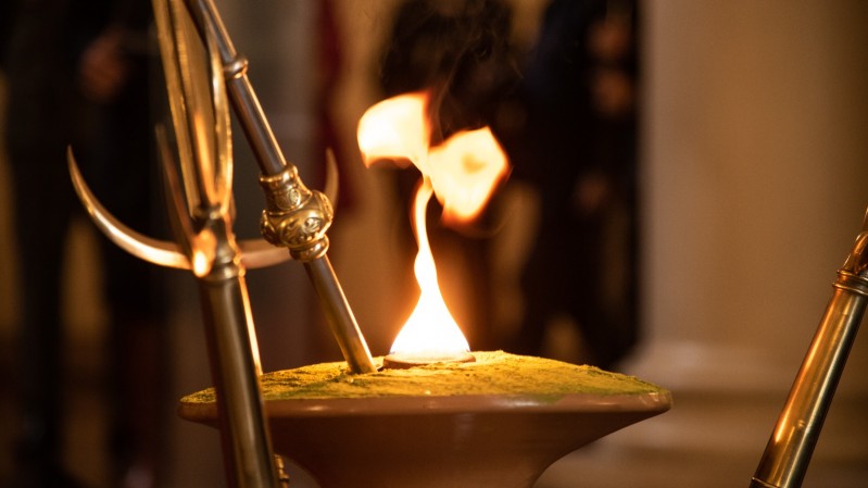 Fotografia em close mostra a Chama Crioula acesa em um recipiente cerimonial de barro. A chama surge no centro da imagem, destacada pela ilumina&ccedil;&atilde;o quente e pelo contraste com o fundo desfocado. Ao redor do recipiente aparecem lan&ccedil;as met&aacute;licas ornamentadas posicionadas verticalmente. O ambiente interno &eacute; iluminado por tons dourados, enquanto pessoas e elementos arquitet&ocirc;nicos ao fundo permanecem fora de foco. 