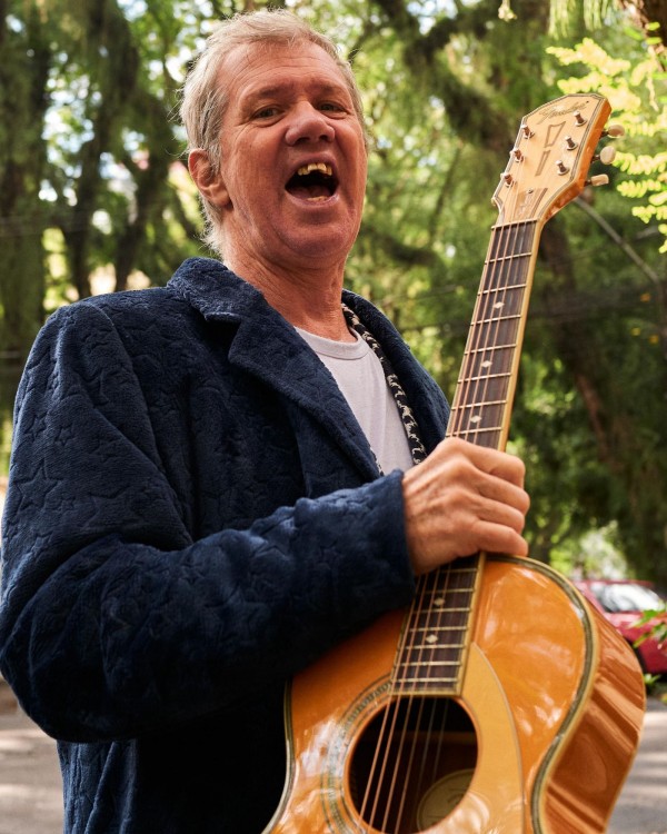 Fotografia em plano aproximado mostra uma pessoa segurando um violão acústico em pé em uma área arborizada. A pessoa veste um casaco azul-escuro de tecido texturizado sobre uma camiseta clara. O instrumento ocupa boa parte do primeiro plano, com destaque para o braço e a caixa de ressonância do violão. Ao fundo, árvores e folhagens aparecem desfocadas, iluminadas por luz natural suave que atravessa a vegetação.