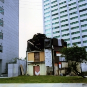 A imagem mostra uma construção pequena e bastante deteriorada, localizada entre dois edifícios altos e modernos. A edificação tem paredes em tons claros, portas e janelas de madeira e partes da cobertura e da estrutura aparentemente danificadas ou inacabadas. À frente, há uma área gramada, uma calçada e parte de uma rua. À direita, aparece uma árvore de copa baixa. Ao fundo, destacam-se os grandes prédios, com muitas janelas e fachadas em tons de branco, cinza e azul. A cena evidencia um forte contraste entre a construção antiga e precária e os edifícios modernos ao redor.