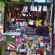 A imagem mostra uma pequena banca ou loja de comércio popular, bastante cheia e organizada com uma grande variedade de mercadorias expostas. O estabelecimento ocupa um espaço compacto, com prateleiras e suportes repletos de produtos do chão ao teto. A cena sugere um ponto de venda de artigos diversos, como materiais escolares, acessórios, itens domésticos e pequenas mercadorias de uso cotidiano.