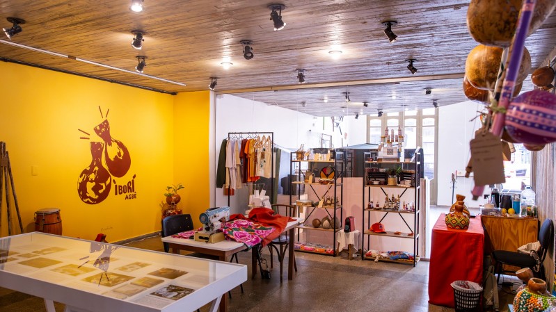 A imagem mostra o interior de uma loja ou espa&ccedil;o expositivo de artesanato e design. O ambiente &eacute; amplo, com piso escuro e teto revestido de madeira, equipado com diversos spots de ilumina&ccedil;&atilde;o direcionada. &Agrave; esquerda, destaca-se uma parede pintada de amarelo intenso, onde h&aacute; um grande logotipo em vermelho com a inscri&ccedil;&atilde;o &ldquo;IBORU ARTE&rdquo; acompanhada de uma ilustra&ccedil;&atilde;o estilizada. Pr&oacute;ximo &agrave; parede, h&aacute; alguns objetos decorativos e um vaso com planta. No primeiro plano, &agrave; esquerda, encontra-se uma mesa branca expositora contendo fotografias, gravuras ou materiais impressos dispostos em fileiras. Ao centro, h&aacute; uma mesa com tecidos coloridos dobrados e outros produtos artesanais. Atr&aacute;s dela, um cabideiro exibe pe&ccedil;as de vestu&aacute;rio em diferentes cores. Mais ao fundo, no centro da imagem, h&aacute; estantes met&aacute;licas com prateleiras de vidro contendo diversos itens decorativos, acess&oacute;rios, cer&acirc;micas, bolsas e objetos artesanais. Grandes janelas na parte posterior permitem a entrada de luz natural. &Agrave; direita, aparecem mais produtos expostos, incluindo recipientes decorativos, pe&ccedil;as artesanais e objetos coloridos pendurados em primeiro plano, parcialmente desfocados, criando sensa&ccedil;&atilde;o de profundidade. Uma mesa coberta por tecido vermelho exibe itens decorativos adicionais.