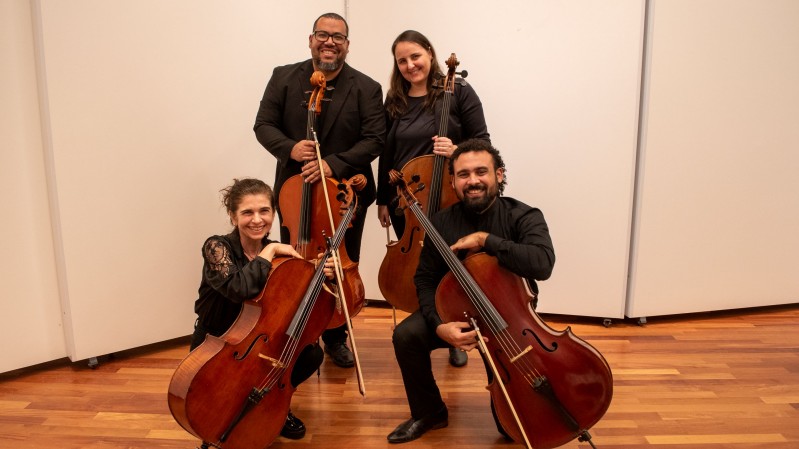 Fotografia de grupo em um ambiente interno com piso de madeira e paredes claras. Quatro m&uacute;sicos vestidos de preto posam lado a lado com instrumentos de cordas de grande porte. Duas pessoas est&atilde;o em p&eacute; ao fundo, cada uma segurando um violoncelo na posi&ccedil;&atilde;o vertical, enquanto duas pessoas est&atilde;o agachadas &agrave; frente, apoiadas em seus instrumentos. Os arcos dos instrumentos aparecem encostados ou apoiados pr&oacute;ximos &agrave;s cordas. A composi&ccedil;&atilde;o &eacute; centralizada, com os m&uacute;sicos ocupando a maior parte da imagem e destacando os tons amadeirados dos violoncelos em contraste com as roupas escuras e o cen&aacute;rio neutro.