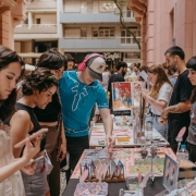 A imagem mostra uma feira ou evento cultural realizado em um espa&ccedil;o aberto, cercado por pr&eacute;dios. Em primeiro plano, &agrave; esquerda, uma mulher de cabelos longos olha para o celular. Ao centro, v&aacute;rias pessoas circulam entre mesas cobertas com livros, revistas, cartazes e outros materiais. Um homem usando bon&eacute;, fones de ouvido e camiseta azul manuseia itens sobre uma das mesas. &Agrave; direita, outras pessoas observam os produtos expostos e conversam. As mesas est&atilde;o organizadas lado a lado, formando corredores de circula&ccedil;&atilde;o, e o ambiente est&aacute; movimentado.