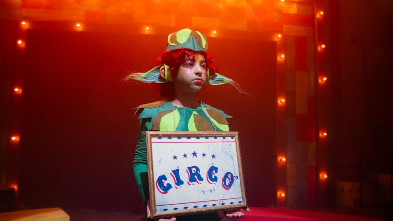 Em um palco iluminado por luzes quentes de camarim distribu&iacute;das ao redor do cen&aacute;rio, uma crian&ccedil;a veste um figurino colorido inspirado no universo circense, com tons de verde, azul e vermelho. No centro da cena, segura uma moldura com a palavra &ldquo;CIRCO&rdquo; escrita em letras decorativas azuis e vermelhas. O fundo apresenta pain&eacute;is coloridos, ilumina&ccedil;&atilde;o c&ecirc;nica e elementos t&iacute;picos de espet&aacute;culo, criando uma atmosfera teatral. A luz avermelhada e alaranjada destaca o figurino e refor&ccedil;a o ambiente de palco.