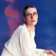 Retrato em ambiente de est&uacute;dio com fundo em tons de azul e rosa que lembram nuvens ao entardecer. Uma pessoa usa vestido longo branco de tecido leve e &oacute;culos de arma&ccedil;&atilde;o redonda. A composi&ccedil;&atilde;o mostra a pessoa agachada, com os bra&ccedil;os apoiados sobre as pernas e o rosto voltado para a c&acirc;mera. A ilumina&ccedil;&atilde;o suave destaca o contraste entre o branco da roupa e as cores vibrantes do cen&aacute;rio.