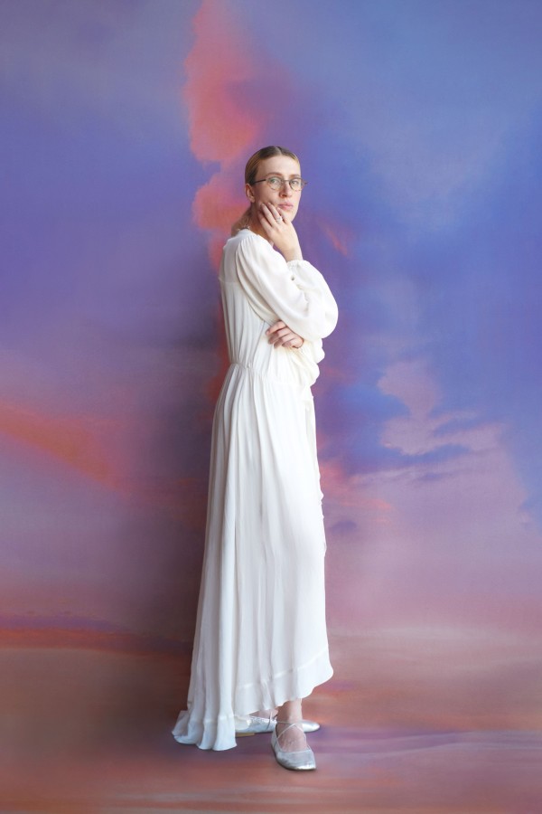 Fotografia vertical em estúdio com cenário desfocado em degradês de azul, lilás e rosa. Ao centro, uma mulher usa óculos de armação redonda e veste um longo vestido branco de mangas compridas e sapatos claros. A pose é feita em pé, com um braço cruzado junto ao corpo e a mão apoiada no rosto.