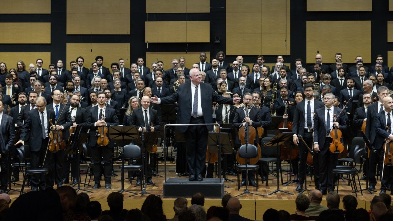 A imagem mostra uma grande orquestra em uma sala de concertos. Ao fundo, h&aacute; um painel com faixas retangulares em tons de madeira e preto. No centro, sobre um pequeno palco, um maestro de terno escuro e cabelos brancos est&aacute; de bra&ccedil;os abertos, conduzindo os m&uacute;sicos. Ao redor dele, dezenas de m&uacute;sicos est&atilde;o posicionados, muitos segurando instrumentos de cordas, como violinos e violoncelos. &Agrave; frente, h&aacute; cadeiras, estantes para partituras e instrumentos. Na parte inferior da imagem, aparecem as silhuetas de pessoas da plateia.