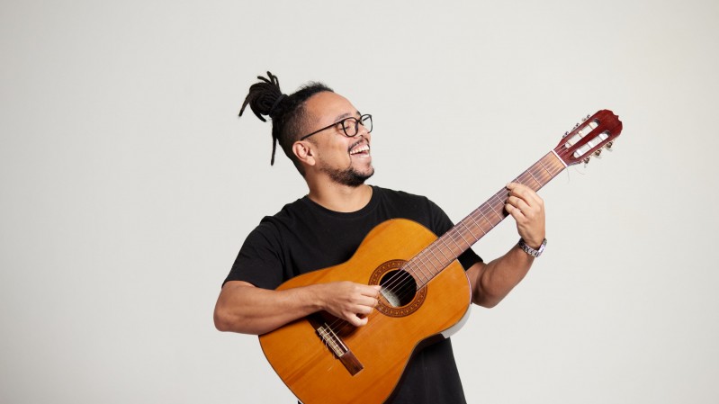 A imagem tem fundo liso na cor cinza-claro. No centro est&aacute; o cantor Rodrigo Fontoura, um homem sorrindo e segurando um viol&atilde;o de madeira. Ele veste uma camiseta preta, usa &oacute;culos de arma&ccedil;&atilde;o escura e tem os cabelos presos em um coque no alto da cabe&ccedil;a. Ele olha para cima e para a direita, enquanto segura o bra&ccedil;o do viol&atilde;o com a m&atilde;o esquerda e posiciona a m&atilde;o direita sobre as cordas, como se estivesse tocando o instrumento. O viol&atilde;o ocupa a parte central e inferior da imagem, com o bra&ccedil;o inclinado para o canto superior direito.
