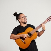 A imagem tem fundo liso na cor cinza-claro. No centro est&aacute; o cantor Rodrigo Fontoura, um homem sorrindo e segurando um viol&atilde;o de madeira. Ele veste uma camiseta preta, usa &oacute;culos de arma&ccedil;&atilde;o escura e tem os cabelos presos em um coque no alto da cabe&ccedil;a. Ele olha para cima e para a direita, enquanto segura o bra&ccedil;o do viol&atilde;o com a m&atilde;o esquerda e posiciona a m&atilde;o direita sobre as cordas, como se estivesse tocando o instrumento. O viol&atilde;o ocupa a parte central e inferior da imagem, com o bra&ccedil;o inclinado para o canto superior direito.