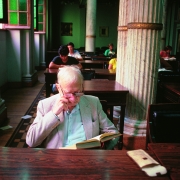 A imagem mostra um ambiente amplo da BPE, iluminado por luz natural. No primeiro plano, ao centro, o poeta Mario Quintana l&ecirc; um livro. Ele &eacute; um homem idoso, de cabelos brancos e &oacute;culos, veste um palet&oacute; claro e l&ecirc; um livro aberto sobre a mesa. Quintana apoia uma das m&atilde;os no rosto enquanto acompanha a leitura. Ao fundo, outras pessoas est&atilde;o sentadas em mesas, lendo ou estudando. O espa&ccedil;o tem grandes colunas decorativas, paredes em tons esverdeados, janelas e m&oacute;veis de madeira escura.
