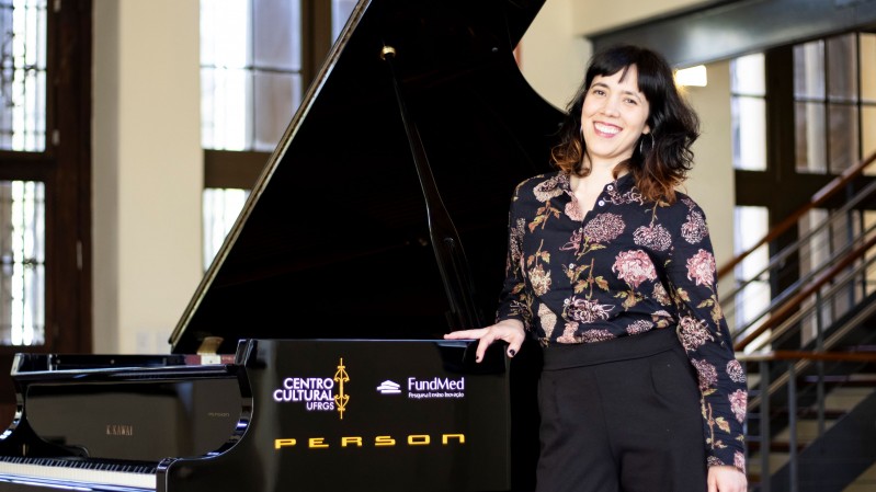 A imagem mostra uma mulher sorrindo, ao lado de um piano de cauda preto, em um ambiente amplo e iluminado. Ela tem cabelos escuros ondulados, usa uma blusa preta estampada com flores e cal&ccedil;a preta, e apoia uma das m&atilde;os sobre o piano. No instrumento, aparecem os logotipos do Centro Cultural e da FundMed, al&eacute;m da marca Person. Ao fundo, h&aacute; janelas altas, colunas claras e uma escada com corrim&atilde;o escuro.
