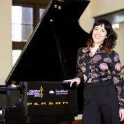 A imagem mostra uma mulher sorrindo, ao lado de um piano de cauda preto, em um ambiente amplo e iluminado. Ela tem cabelos escuros ondulados, usa uma blusa preta estampada com flores e cal&ccedil;a preta, e apoia uma das m&atilde;os sobre o piano. No instrumento, aparecem os logotipos do Centro Cultural e da FundMed, al&eacute;m da marca Person. Ao fundo, h&aacute; janelas altas, colunas claras e uma escada com corrim&atilde;o escuro.
