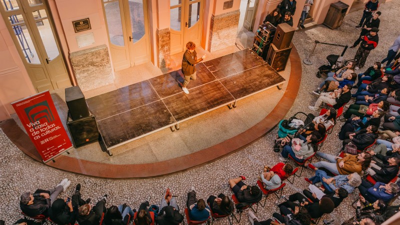 A imagem mostra um evento realizado no sagu&atilde;o de um pr&eacute;dio hist&oacute;rico, com piso de mosaico e paredes em tons claros, detalhes em m&aacute;rmore e portas de madeira. No centro, h&aacute; um pequeno palco de madeira, onde uma pessoa est&aacute; em p&eacute;, ao lado de equipamentos de som. O p&uacute;blico est&aacute; sentado e reunido ao redor do palco, acompanhando a apresenta&ccedil;&atilde;o. &Agrave; esquerda, h&aacute; um painel vermelho com informa&ccedil;&otilde;es sobre o evento.