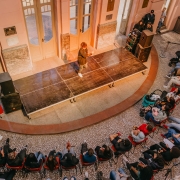 A imagem mostra um evento realizado no sagu&atilde;o de um pr&eacute;dio hist&oacute;rico, com piso de mosaico e paredes em tons claros, detalhes em m&aacute;rmore e portas de madeira. No centro, h&aacute; um pequeno palco de madeira, onde uma pessoa est&aacute; em p&eacute;, ao lado de equipamentos de som. O p&uacute;blico est&aacute; sentado e reunido ao redor do palco, acompanhando a apresenta&ccedil;&atilde;o. &Agrave; esquerda, h&aacute; um painel vermelho com informa&ccedil;&otilde;es sobre o evento.