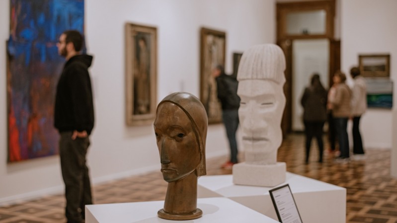 A imagem mostra uma sala de exposi&ccedil;&atilde;o de arte do MARGS, com paredes brancas e piso de madeira. Em primeiro plano, sobre pedestais brancos, h&aacute; duas esculturas de cabe&ccedil;as: uma menor, de aspecto met&aacute;lico e em tom marrom, e outra maior, branca, com tra&ccedil;os estilizados. Ao fundo, pessoas circulam e observam as obras expostas nas paredes. &Agrave; esquerda, h&aacute; uma grande pintura colorida, enquanto outras obras emolduradas est&atilde;o distribu&iacute;das pelas paredes.