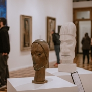 A imagem mostra uma sala de exposi&ccedil;&atilde;o de arte do MARGS, com paredes brancas e piso de madeira. Em primeiro plano, sobre pedestais brancos, h&aacute; duas esculturas de cabe&ccedil;as: uma menor, de aspecto met&aacute;lico e em tom marrom, e outra maior, branca, com tra&ccedil;os estilizados. Ao fundo, pessoas circulam e observam as obras expostas nas paredes. &Agrave; esquerda, h&aacute; uma grande pintura colorida, enquanto outras obras emolduradas est&atilde;o distribu&iacute;das pelas paredes.