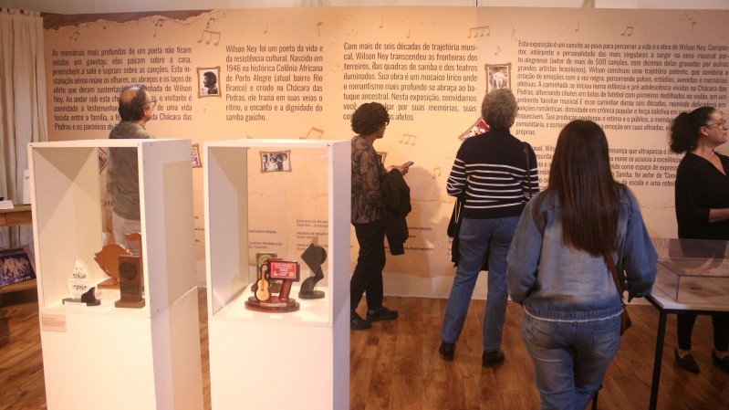 A imagem mostra uma parte da exposi&ccedil;&atilde;o &ldquo;Mem&oacute;ria Cantada: Wilson Ney&rdquo;. A sala tem as paredes escritas com a traj&eacute;toria do artista e algumas pessoas circulam pelo local. 