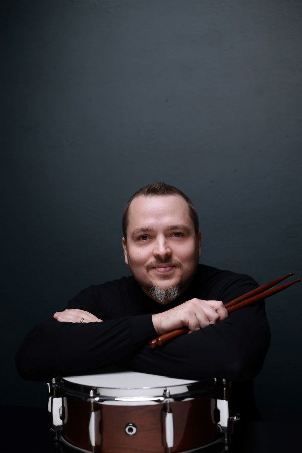Retrato em estúdio com fundo azul-acinzentado mostra Douglas Gutjahr apoiando os braços sobre uma caixa clara e segurando duas baquetas de percussão. À frente, aparece um tambor de madeira com pele branca, parcialmente visível no primeiro plano. Douglas veste roupa preta de mangas longas e está posicionada de frente para a câmera. A iluminação suave destaca o rosto, as baquetas e os detalhes do instrumento, enquanto o fundo uniforme concentra a atenção na composição do retrato.