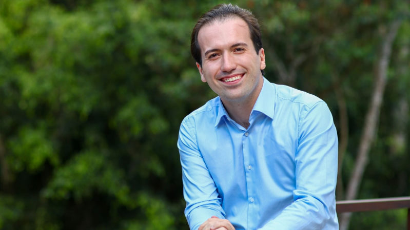 José Soares est&aacute; apoiado em um guarda-corpo met&aacute;lico em uma &aacute;rea externa cercada por vegeta&ccedil;&atilde;o. Vestindo camisa azul-clara e cal&ccedil;a escura, est&aacute; com uma m&atilde;o sobre a outra enquanto permanece voltado para a c&acirc;mera. Ao fundo, &aacute;rvores e folhagens desfocadas formam uma paisagem verde que ocupa toda a composi&ccedil;&atilde;o. A luz natural ilumina a cena e destaca os tons suaves da roupa em contraste com o ambiente ao ar livre.
