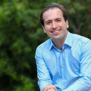 José Soares est&aacute; apoiado em um guarda-corpo met&aacute;lico em uma &aacute;rea externa cercada por vegeta&ccedil;&atilde;o. Vestindo camisa azul-clara e cal&ccedil;a escura, est&aacute; com uma m&atilde;o sobre a outra enquanto permanece voltado para a c&acirc;mera. Ao fundo, &aacute;rvores e folhagens desfocadas formam uma paisagem verde que ocupa toda a composi&ccedil;&atilde;o. A luz natural ilumina a cena e destaca os tons suaves da roupa em contraste com o ambiente ao ar livre.