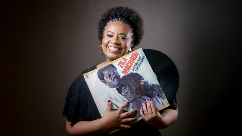 Imagem mostra a cantora Marguerite Ramos Silva, uma mulher negra, sorrindo, em frente a um fundo escuro. Ela tem cabelos crespos curtos, usa blusa preta e segura &agrave; frente do corpo um disco de vinil e a capa de um &aacute;lbum. Na capa, aparecem fotografias de pessoas e o t&iacute;tulo &ldquo;ZAN&hellip; MAR&hellip;&rdquo; em letras vermelhas. A mulher olha para a c&acirc;mera enquanto segura os objetos com as duas m&atilde;os.