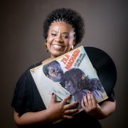 Imagem mostra a cantora Marguerite Ramos Silva, uma mulher negra, sorrindo, em frente a um fundo escuro. Ela tem cabelos crespos curtos, usa blusa preta e segura &agrave; frente do corpo um disco de vinil e a capa de um &aacute;lbum. Na capa, aparecem fotografias de pessoas e o t&iacute;tulo &ldquo;ZAN&hellip; MAR&hellip;&rdquo; em letras vermelhas. A mulher olha para a c&acirc;mera enquanto segura os objetos com as duas m&atilde;os.