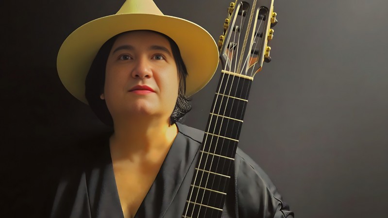 A imagem mostra a violinista Andrea Perrone. Ela usa um chap&eacute;u de aba larga em tom bege e uma roupa preta. Ela segura um viol&atilde;o, que aparece em destaque no lado direito da imagem, com o bra&ccedil;o e as cordas vis&iacute;veis. Ao fundo, h&aacute; um cen&aacute;rio escuro e uniforme, que destaca o rosto, o chap&eacute;u e o instrumento.