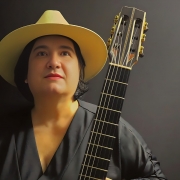 A imagem mostra a violinista Andrea Perrone. Ela usa um chap&eacute;u de aba larga em tom bege e uma roupa preta. Ela segura um viol&atilde;o, que aparece em destaque no lado direito da imagem, com o bra&ccedil;o e as cordas vis&iacute;veis. Ao fundo, h&aacute; um cen&aacute;rio escuro e uniforme, que destaca o rosto, o chap&eacute;u e o instrumento.