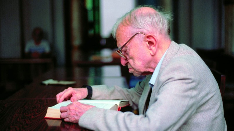 A imagem mostra o poeta e escritor Mario Quintana sentado &agrave; mesa, concentrado na leitura de um livro aberto. Ele est&aacute; posicionado de perfil, voltado para a esquerda da imagem, usando &oacute;culos e vestindo um palet&oacute; claro sobre uma camisa de tom suave. O cen&aacute;rio parece ser um ambiente interno, possivelmente uma biblioteca, sala de estudos ou espa&ccedil;o de leitura. H&aacute; uma mesa longa de madeira escura ocupando boa parte do primeiro plano. Ao fundo, o ambiente est&aacute; desfocado, com colunas ou estruturas verticais vis&iacute;veis e outra pessoa distante, tamb&eacute;m desfocada.