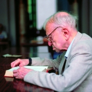 A imagem mostra o poeta e escritor Mario Quintana sentado &agrave; mesa, concentrado na leitura de um livro aberto. Ele est&aacute; posicionado de perfil, voltado para a esquerda da imagem, usando &oacute;culos e vestindo um palet&oacute; claro sobre uma camisa de tom suave. O cen&aacute;rio parece ser um ambiente interno, possivelmente uma biblioteca, sala de estudos ou espa&ccedil;o de leitura. H&aacute; uma mesa longa de madeira escura ocupando boa parte do primeiro plano. Ao fundo, o ambiente est&aacute; desfocado, com colunas ou estruturas verticais vis&iacute;veis e outra pessoa distante, tamb&eacute;m desfocada.